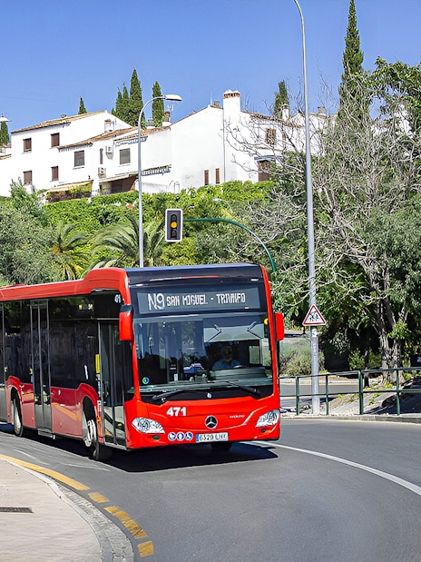 Granada city bus on route near Alsa station, surrounded by greenery and buildings.