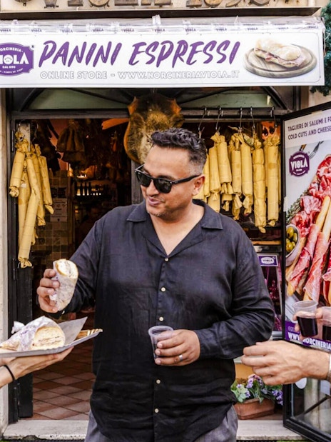 Guide serving panini to guests at a Rome food tour outside a deli.