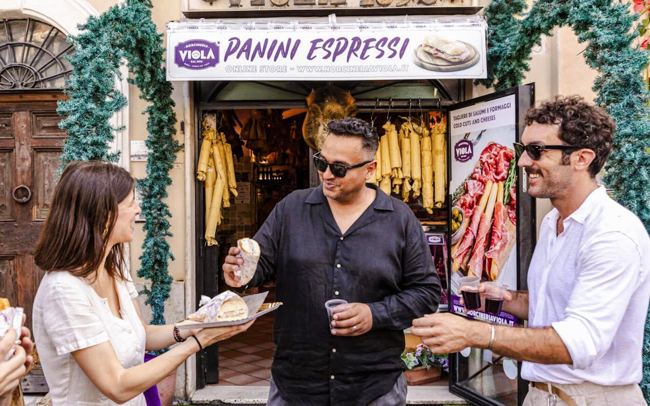 Guide serving panini to guests at a Rome food tour outside a deli.