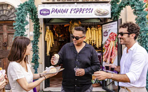 Guide serving panini to guests at a Rome food tour outside a deli.