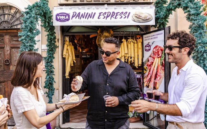 Guide serving panini to guests at a Rome food tour outside a deli.