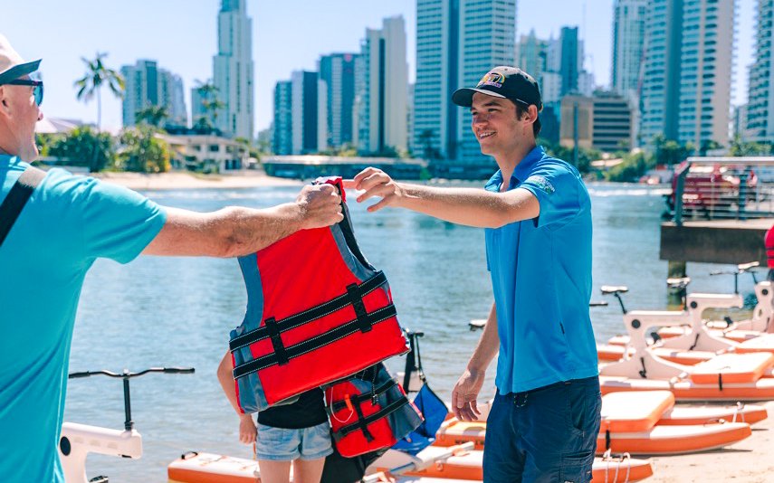 Guide handing life jacket to tourist by the waterfront in a cityscape setting.