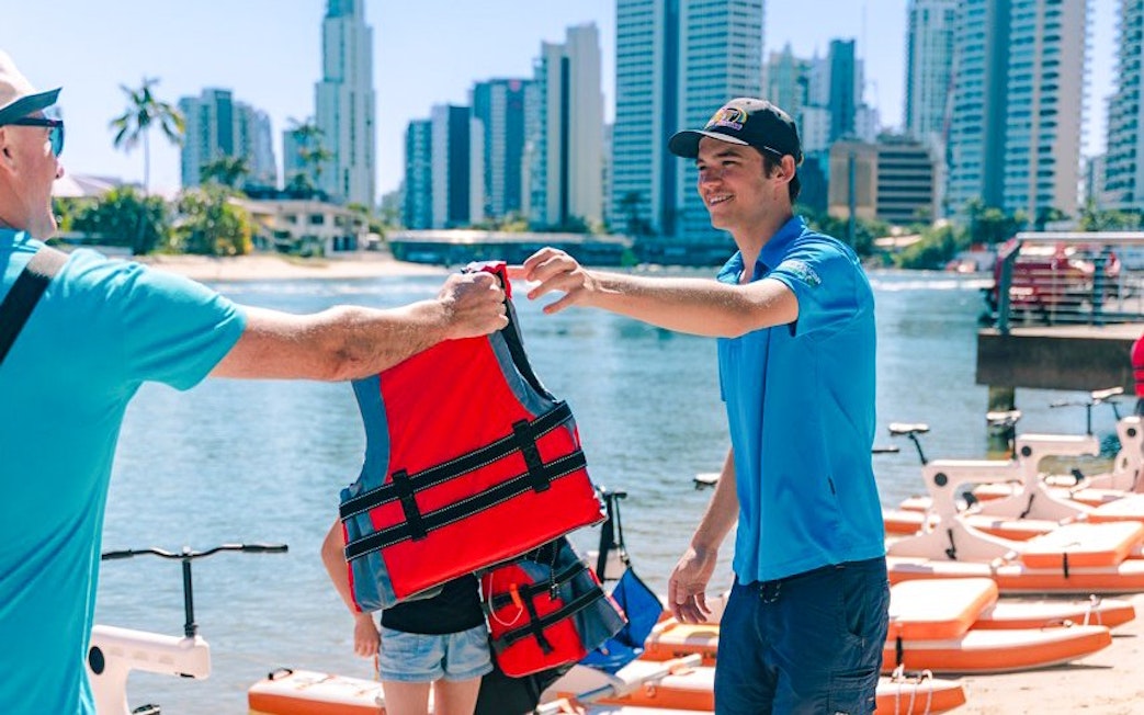 Guide handing life jacket to tourist by the waterfront in a cityscape setting.