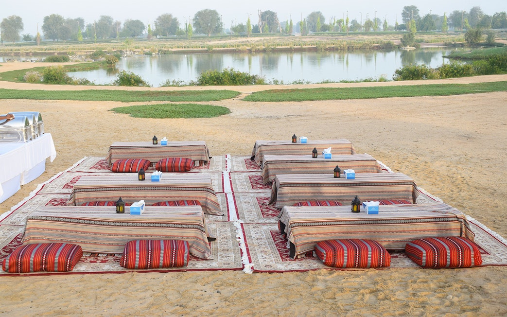 Mattresses and tables set up for guests at a desert safari, with a lake view in the background.