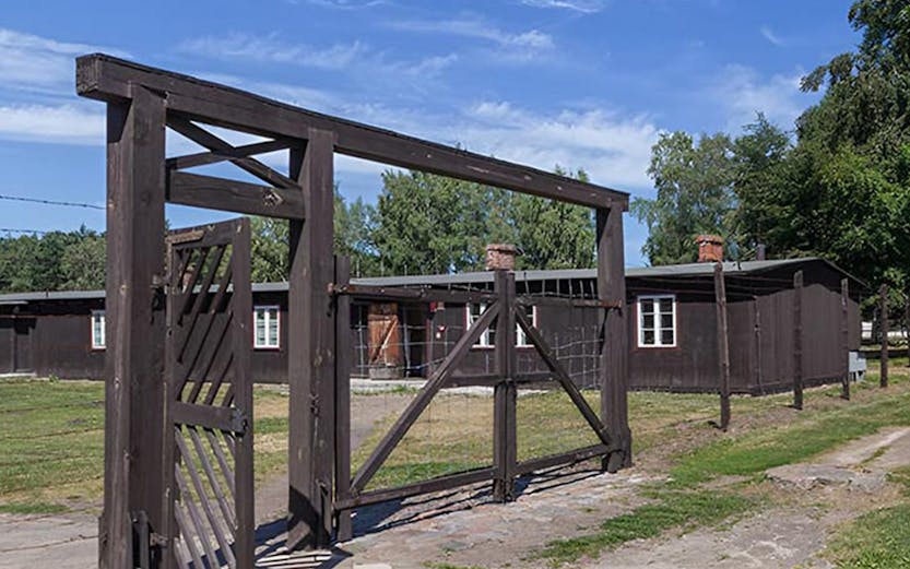 Entrance gate of Stutthof Concentration Camp with wooden barracks in the background.