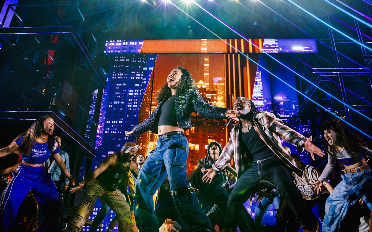 Performers dancing energetically on stage with a vibrant cityscape backdrop in Hell's Kitchen.