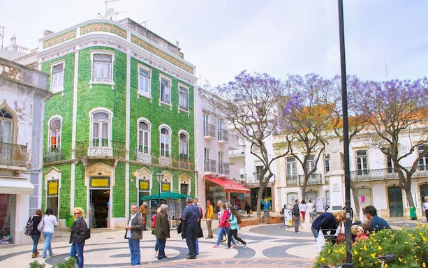 People walking in a historic square with a green-tiled building in Western Algarve.
