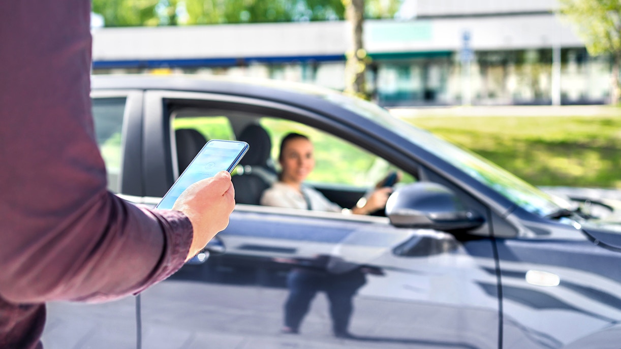 Person using smartphone for shared transfer service with car and driver waiting.