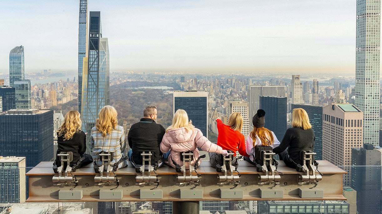 People sitting on a beam at Top of the Rock, Rockefeller Center, New York City skyline in background.