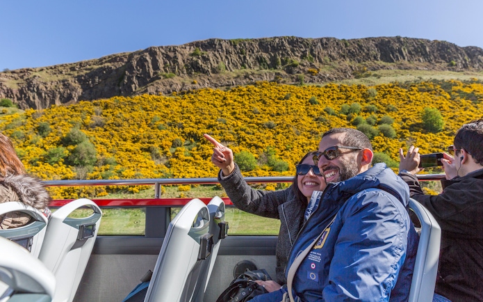 Passengers enjoying the view from the upper deck of a HOHO bus near a hillside with yellow flowers.