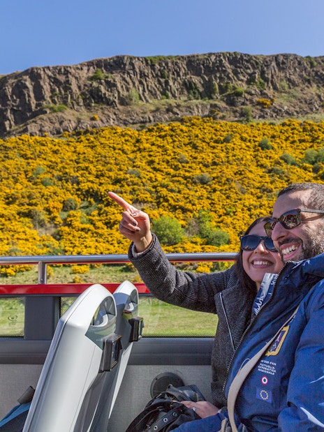 Passengers enjoying the view from the upper deck of a HOHO bus near a hillside with yellow flowers.