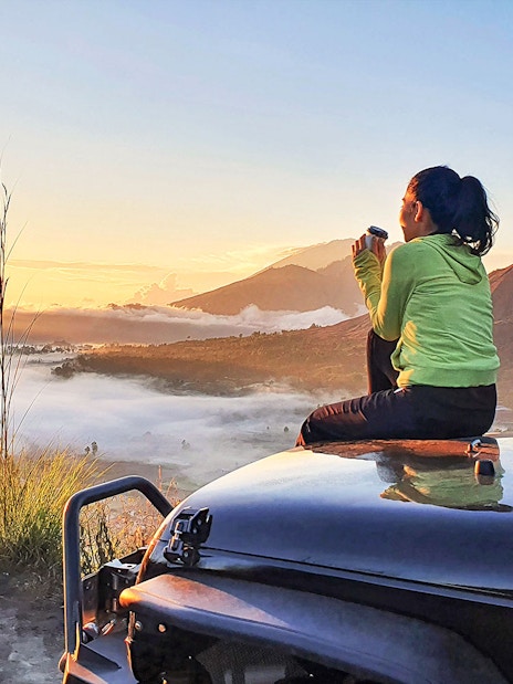 Woman sitting on car hood enjoying sunrise view of Mount Batur, Bali.
