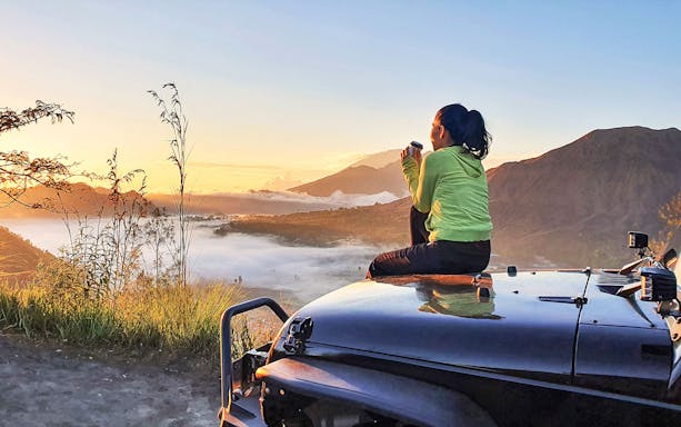 Woman sitting on car hood enjoying sunrise view of Mount Batur, Bali.