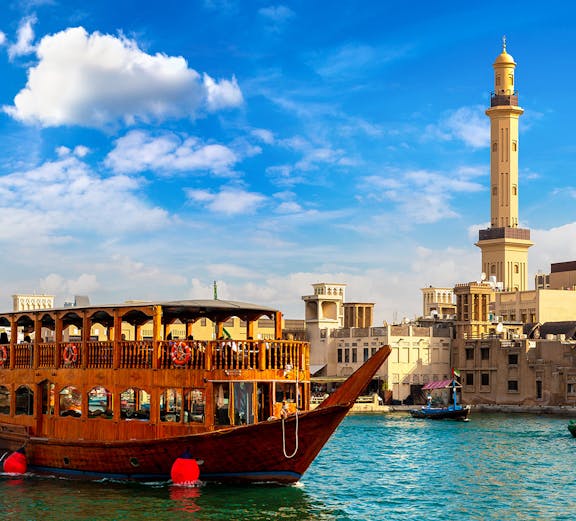 Traditional dhow boat sailing on Dubai Creek with city skyline in the background.