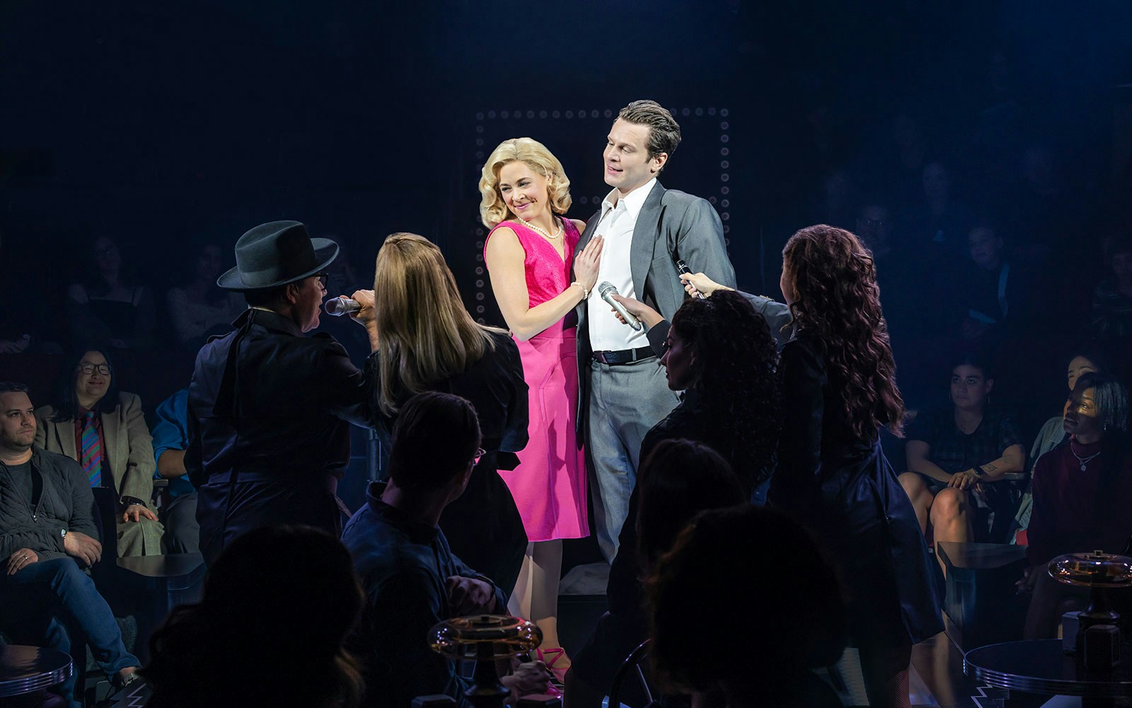Performers on stage during a live theater show with audience in the background.