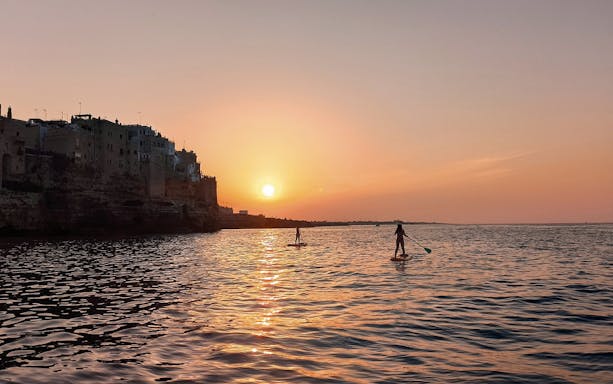 Paddleboarders at sunset near Polignano a Mare coastline.