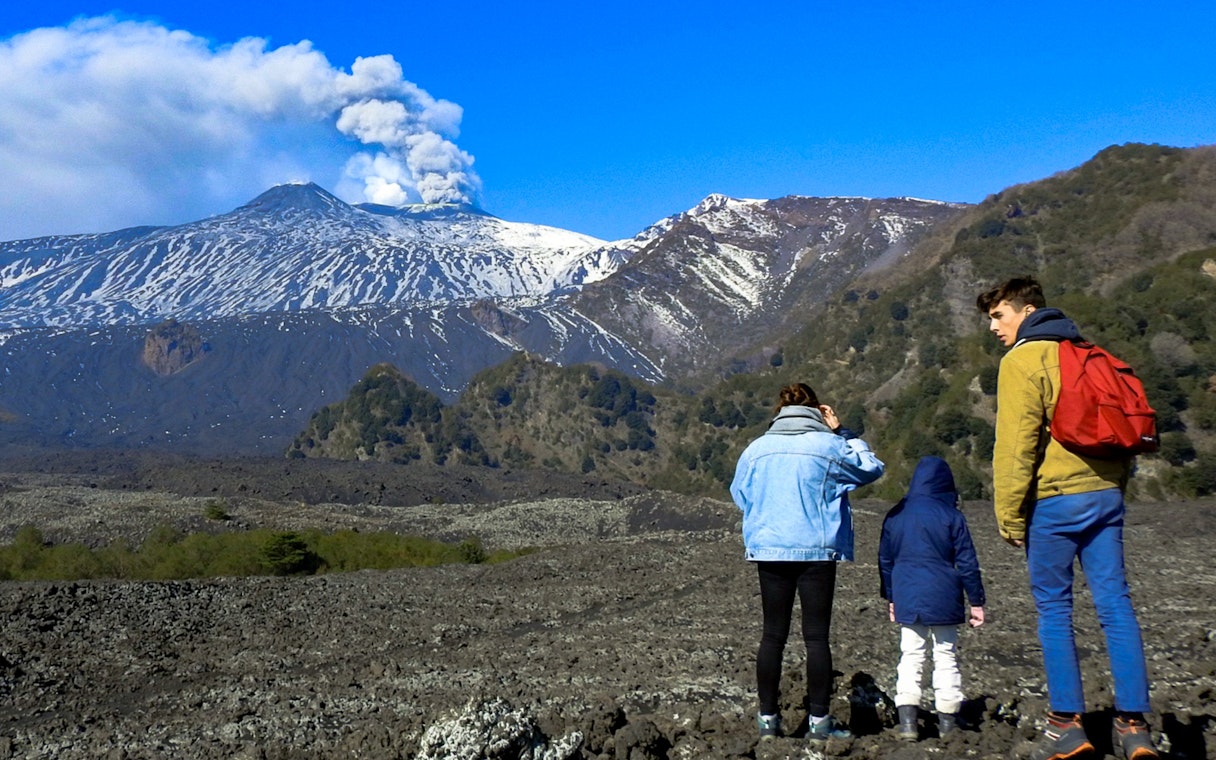 Hikers exploring volcanic landscape of Mount Etna with smoke rising from the crater.