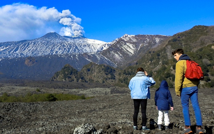 Hikers exploring volcanic landscape of Mount Etna with smoke rising from the crater.