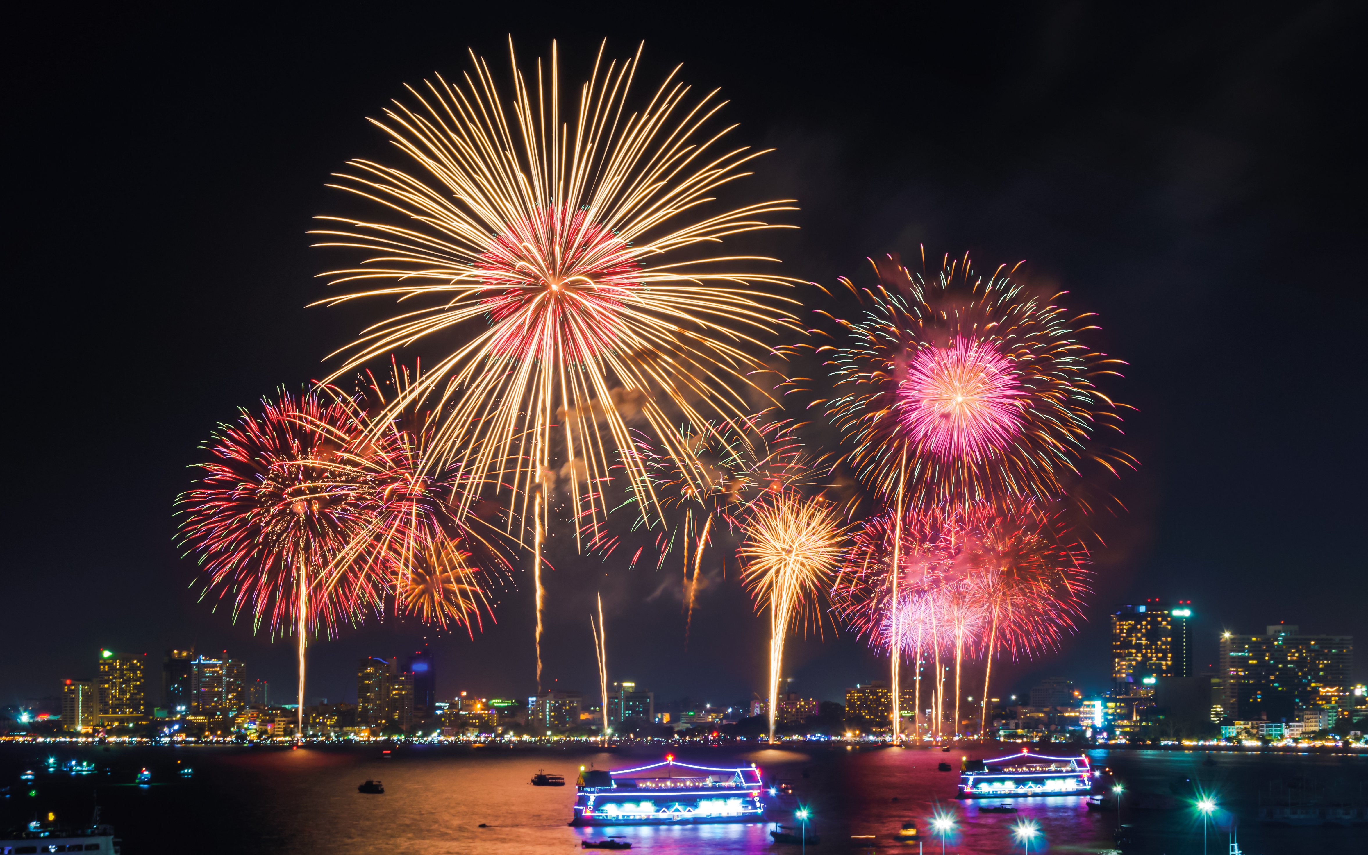 Fireworks display over city skyline with illuminated boats on the water.