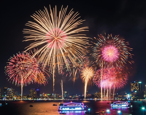 Fireworks display over city skyline with illuminated boats on the water.