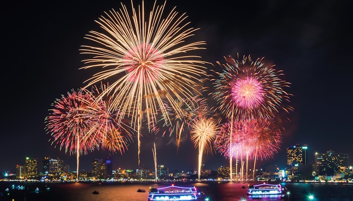 Fireworks display over city skyline with illuminated boats on the water.
