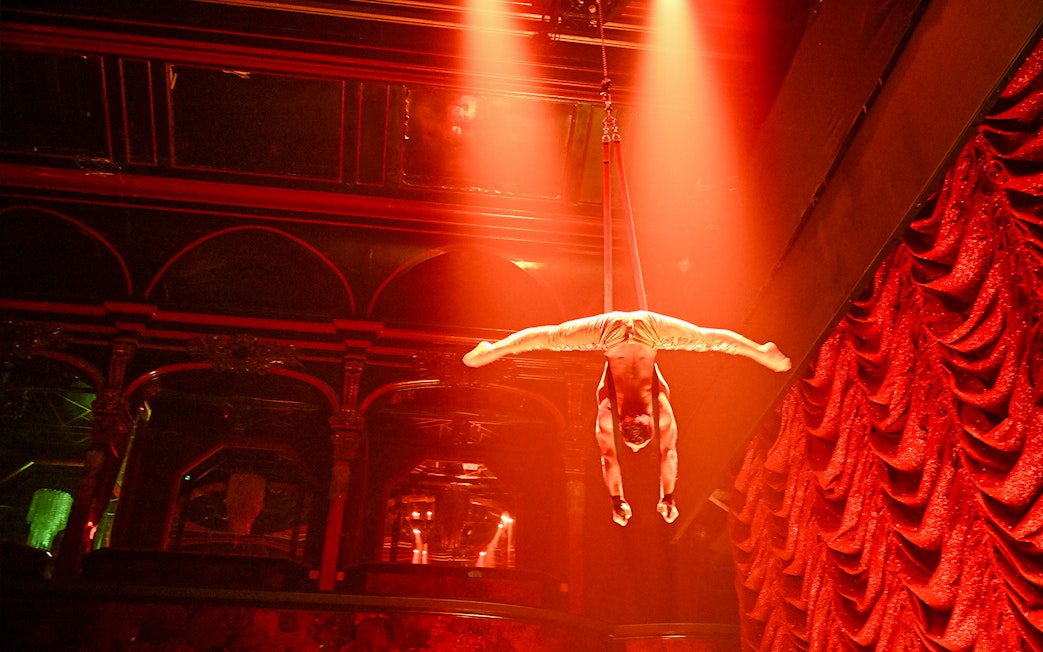 Aerial performer in Paris cabaret show, suspended upside down under red stage lights.