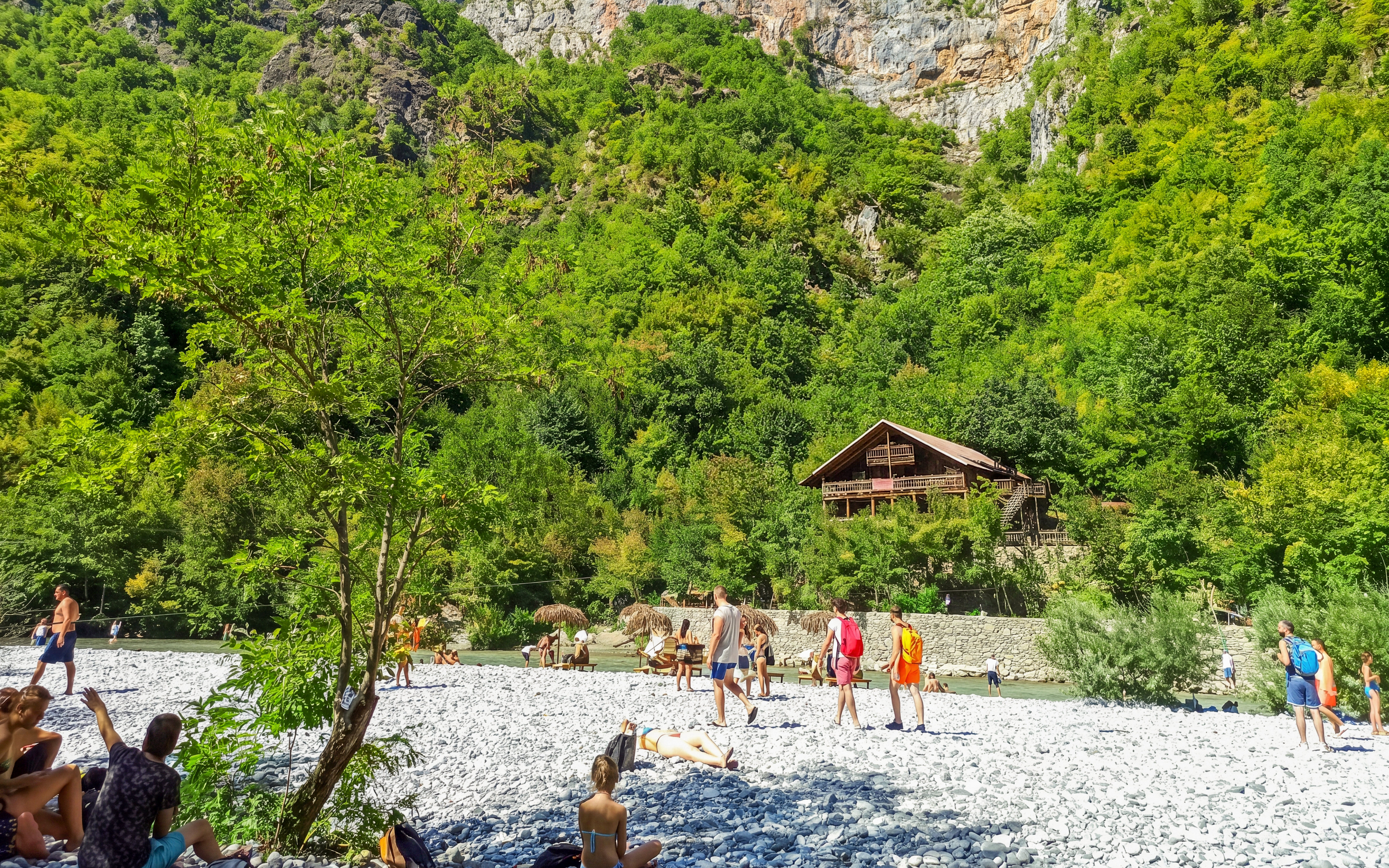 Visitors relaxing by Shala River with a wooden cabin in the lush green landscape, Komani Lake day trip.