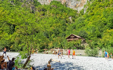 Visitors relaxing by Shala River with a wooden cabin in the lush green landscape, Komani Lake day trip.