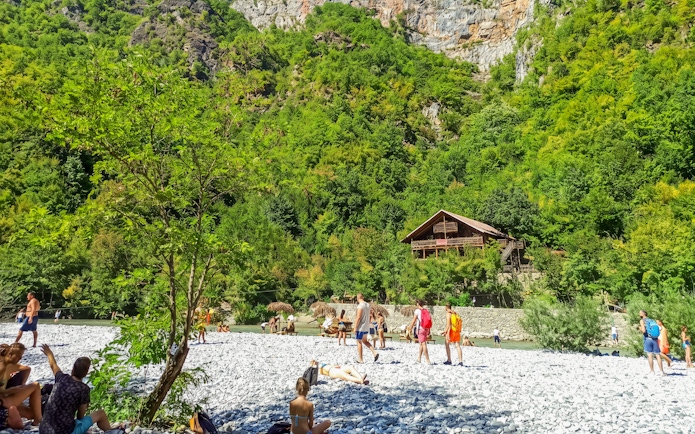 Visitors relaxing by Shala River with a wooden cabin in the lush green landscape, Komani Lake day trip.
