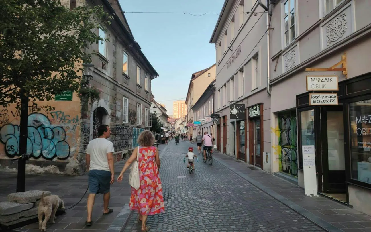 Ljubljana street with people walking and cycling, featuring local shops and graffiti.