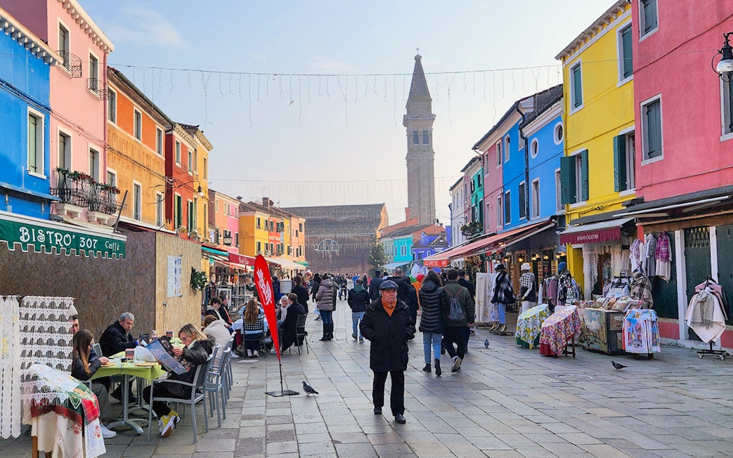 Colorful buildings and bustling street in Burano, Italy with people dining and shopping.