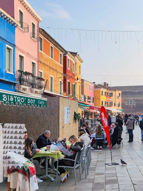 Colorful buildings and bustling street in Burano, Italy with people dining and shopping.