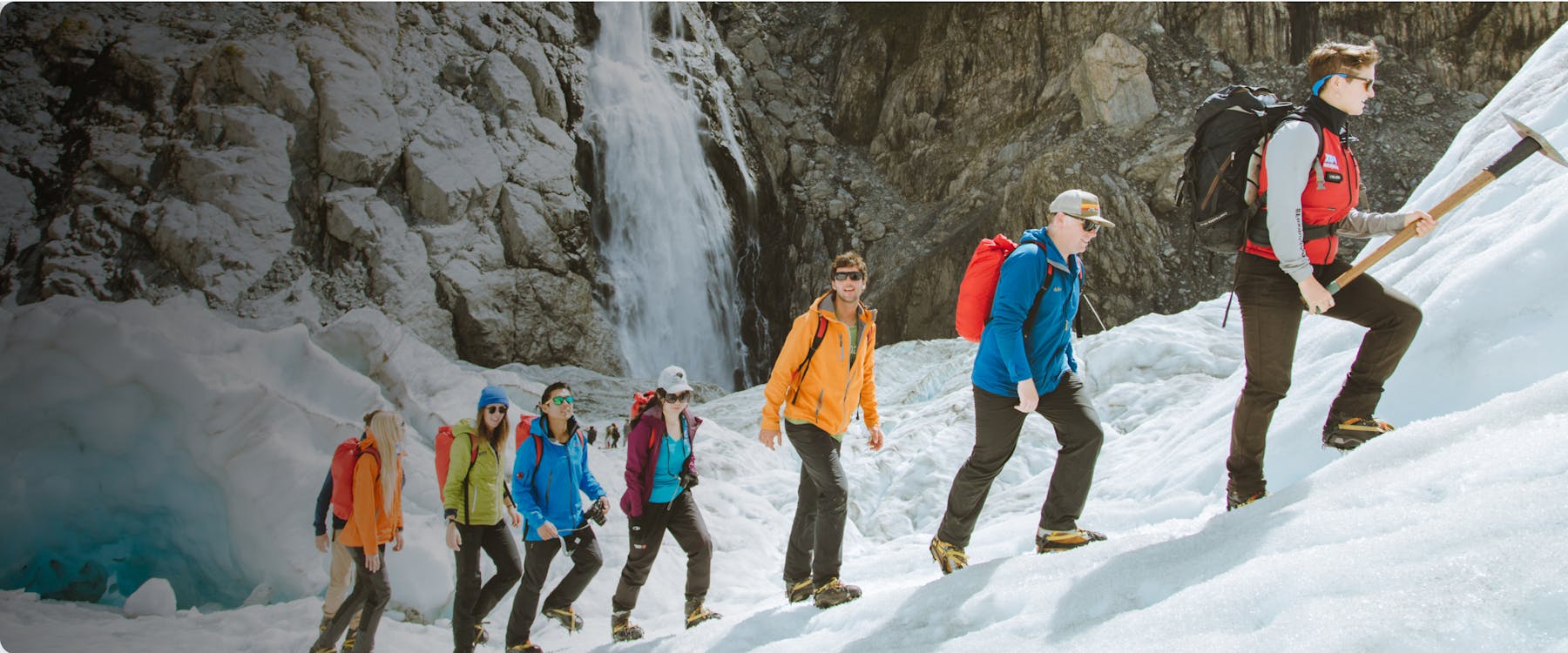 Hikers with gear trekking on Fox Glacier, New Zealand, near a waterfall.