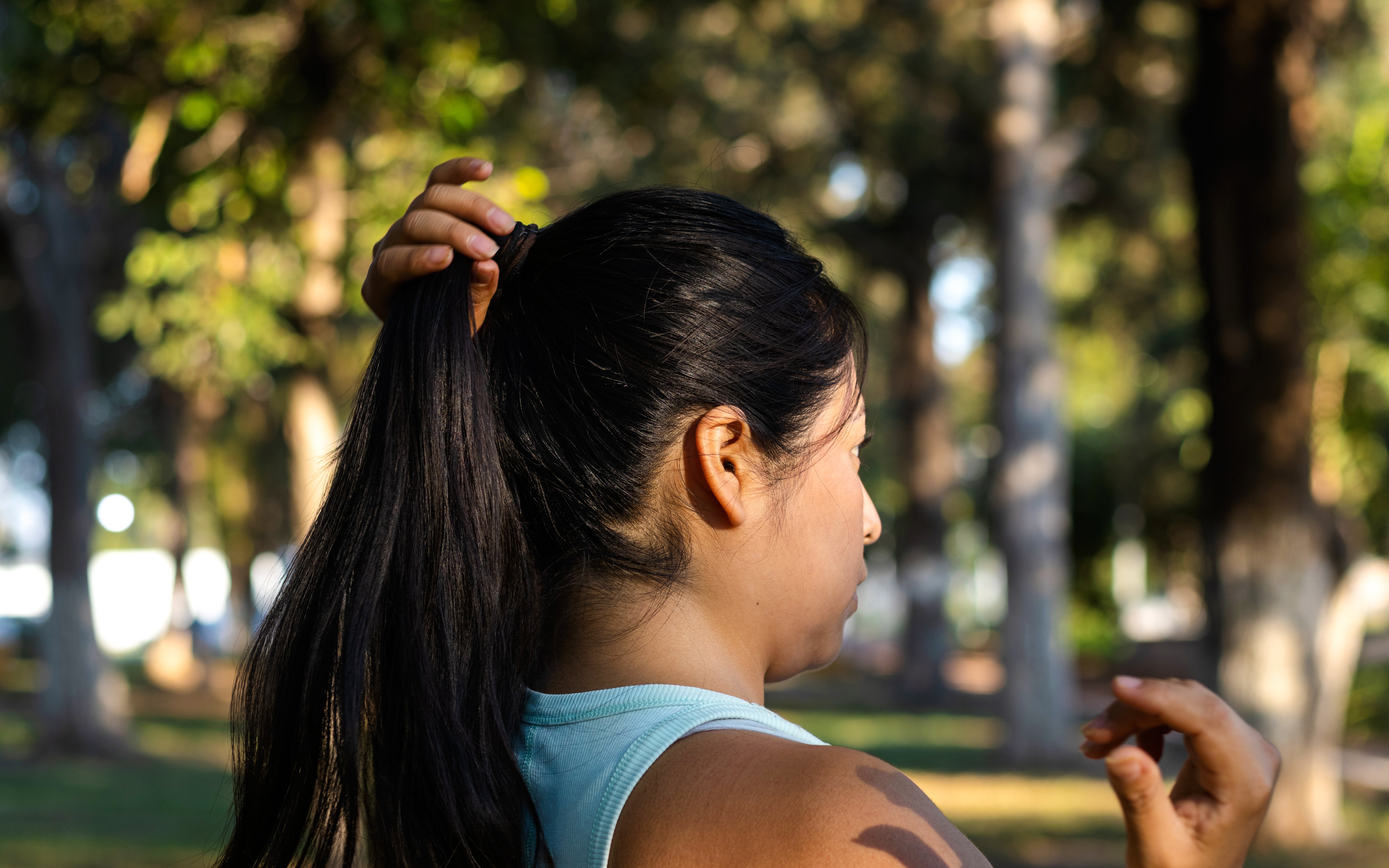 Woman tying long black hair into a ponytail in a park setting.