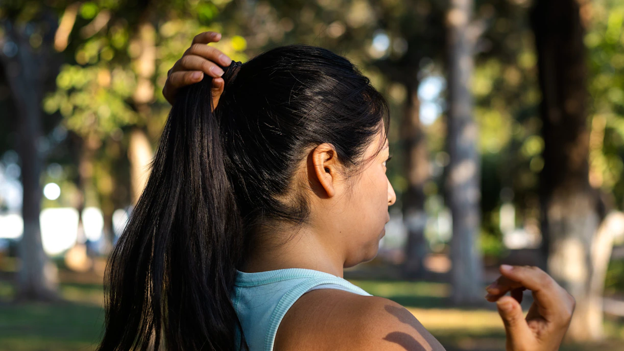 Woman tying long black hair into a ponytail in a park setting.