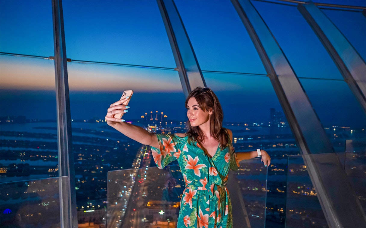 Person taking a selfie at The View At The Palm, Dubai skyline in background.