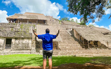 Visitor at Ek Balam pyramid with thatched structures and jungle backdrop in Yucatan, Mexico.