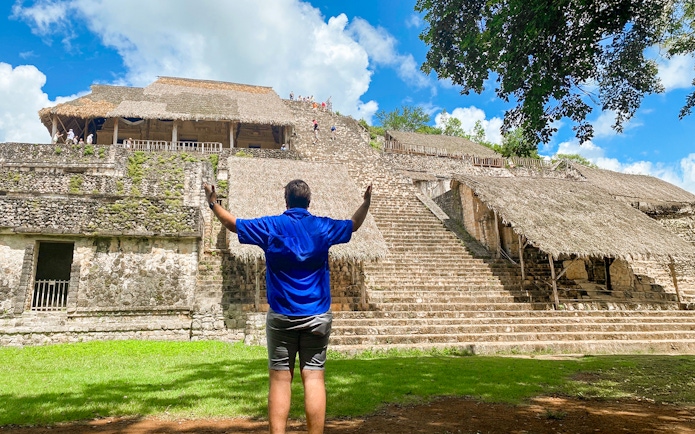 Visitor at Ek Balam pyramid with thatched structures and jungle backdrop in Yucatan, Mexico.