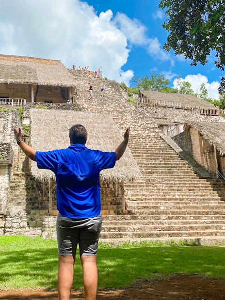 Visitor at Ek Balam pyramid with thatched structures and jungle backdrop in Yucatan, Mexico.