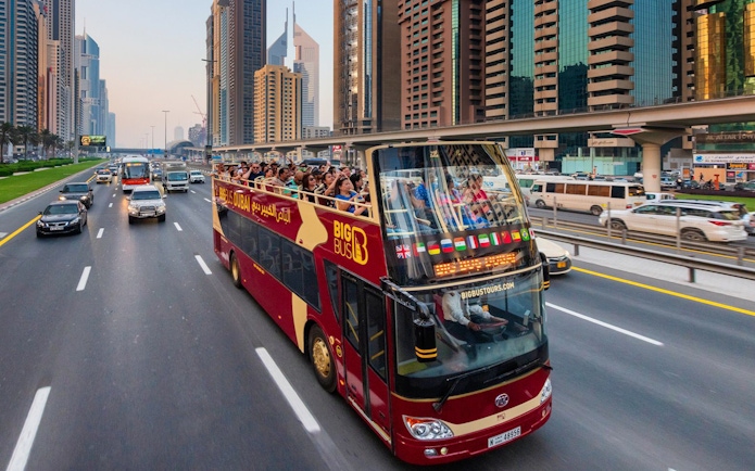 Open-top tour bus on Dubai highway with city skyscrapers in the background.