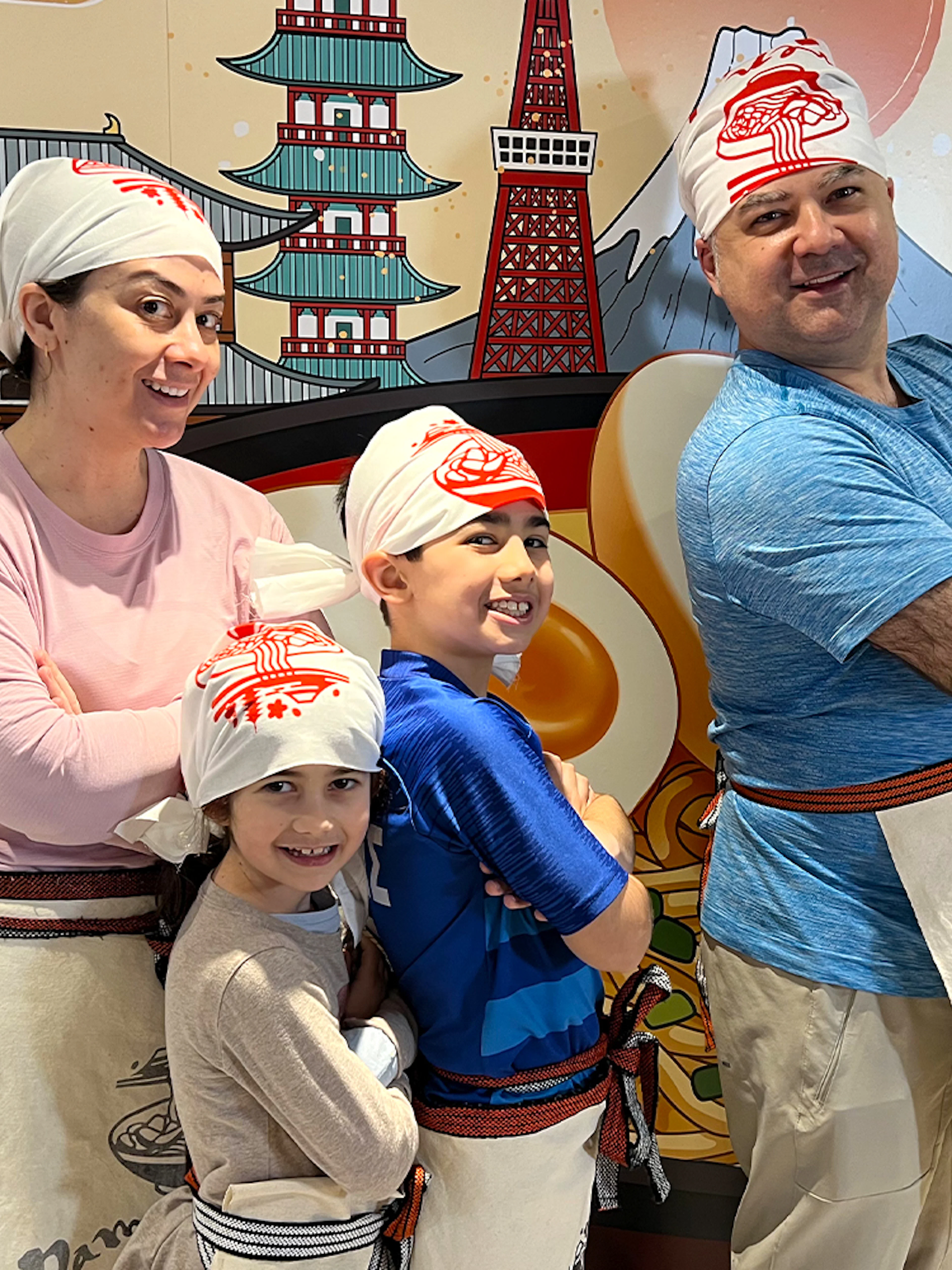 Family posing in front of ramen-themed mural at Ramen Factory Kyoto cooking class.