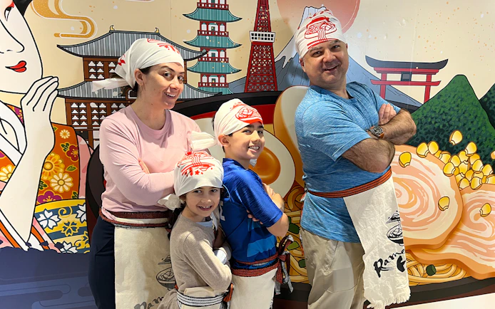 Family posing in front of ramen-themed mural at Ramen Factory Kyoto cooking class.