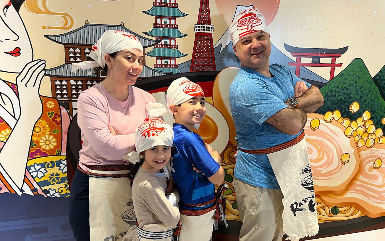 Family posing in front of ramen-themed mural at Ramen Factory Kyoto cooking class.