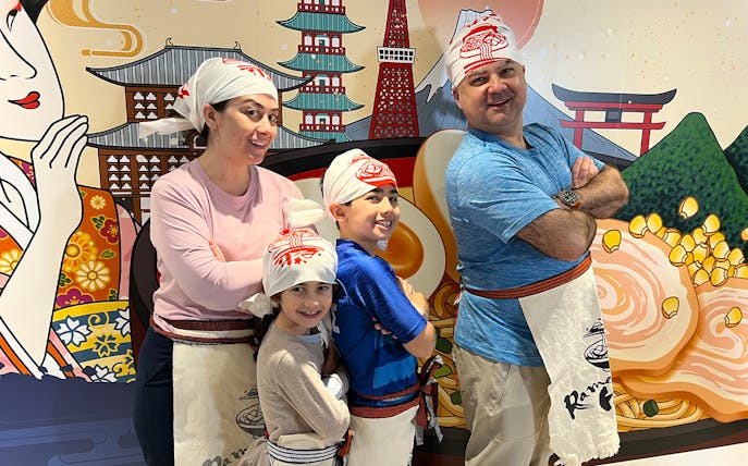 Family posing in front of ramen-themed mural at Ramen Factory Kyoto cooking class.