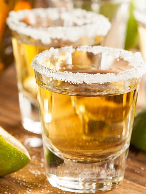 Tequila shots with salt rim and lime wedges on a wooden table.