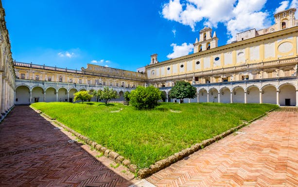 Cloister and gardens of San Martino in Naples with arched walkways and green lawn.