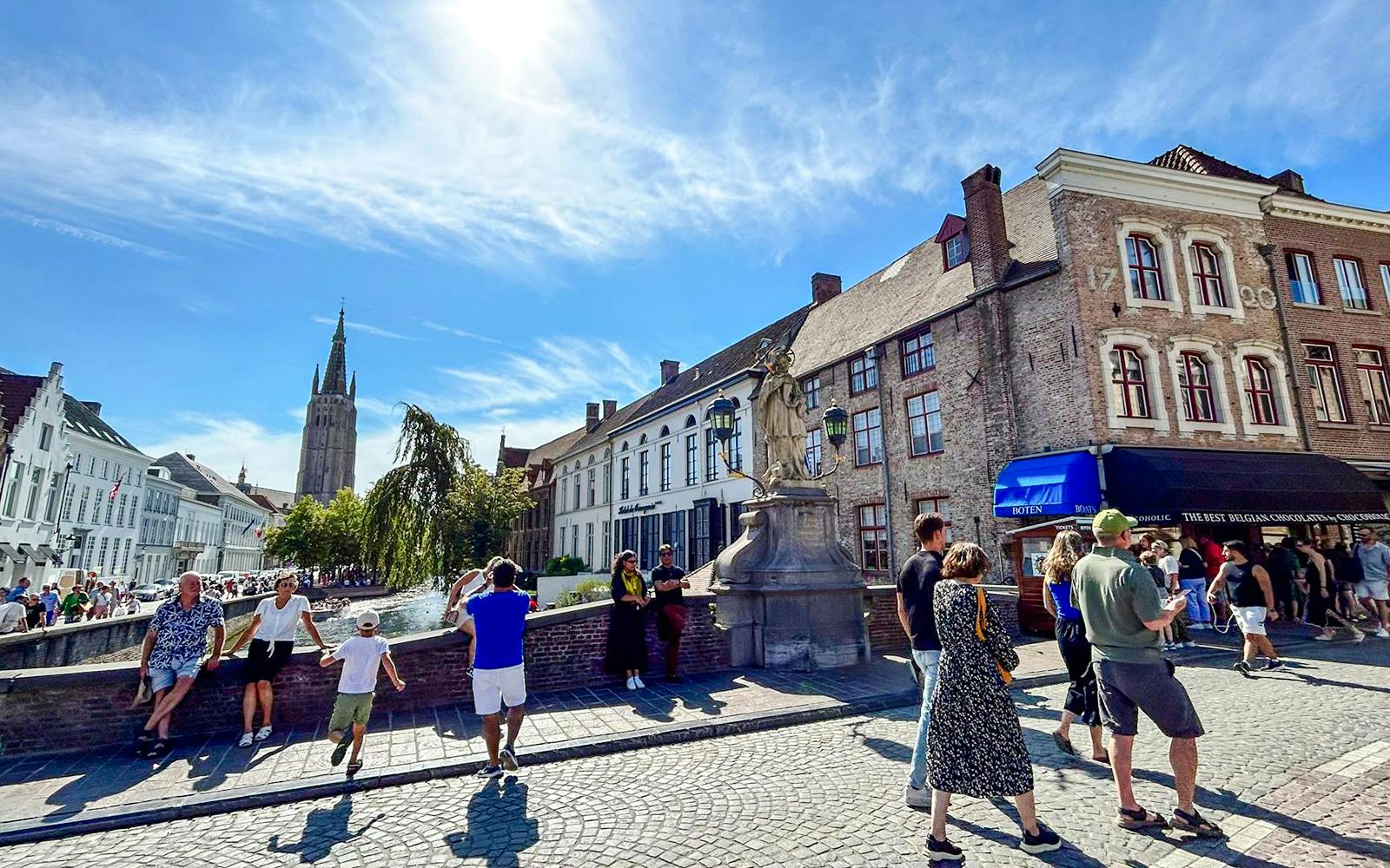 Sunny day in Bruges historic center with people near canal and medieval buildings.