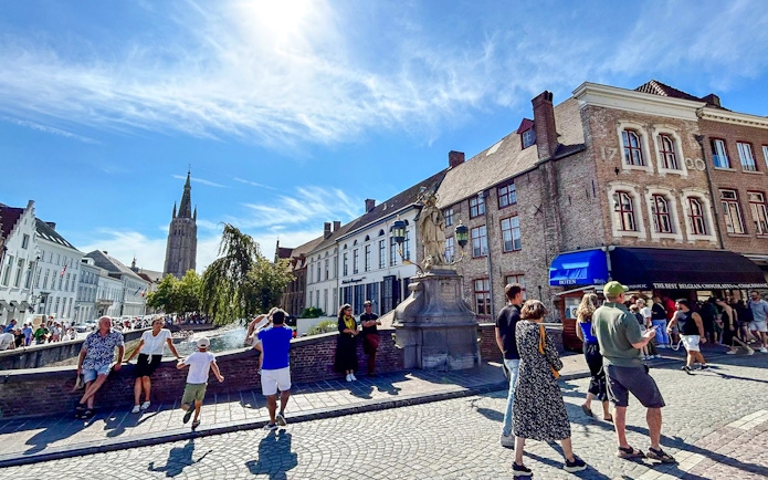 Sunny day in Bruges historic center with people near canal and medieval buildings.