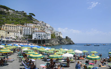 Beachgoers under umbrellas with Amalfi Coast cliffside buildings and boats in the sea.