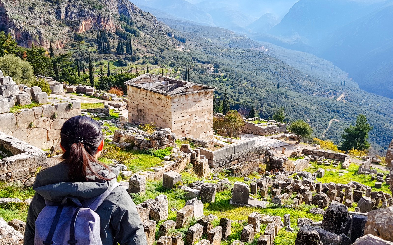 Visitor exploring ancient ruins at Delphi Archaeological Site, Greece, with mountainous landscape.